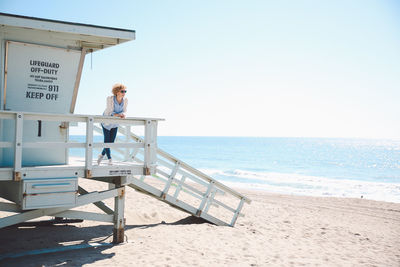 Mid adult woman standing on lifeguard hut at beach against clear sky