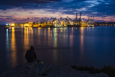 Man sitting on illuminated harbor against sky at dusk