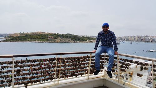 Man sitting on railing with love locks against sky