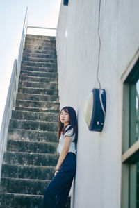 Portrait of young woman standing by wall on stairs at building terrace