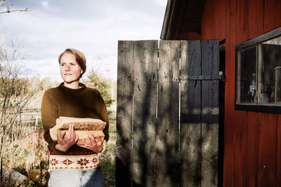 Thoughtful young woman holding firewood while standing outside cottage