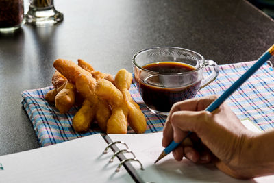 Fried dough stick and cup of black coffee near by woman writing in notebook on the desk