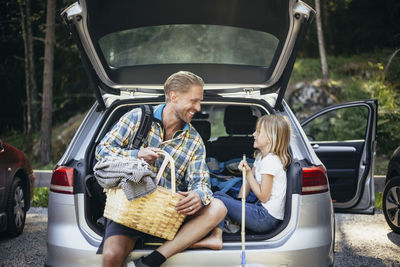 Smiling father with picnic basket talking to daughter while sitting in car trunk