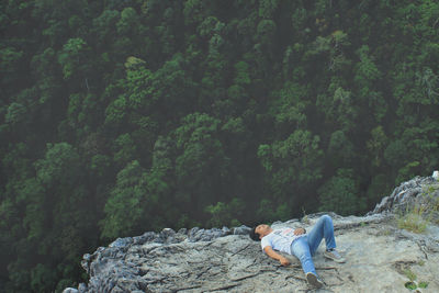 High angle view of young man lying on rocky mountain overlooking dense forest