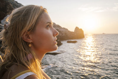 Side view of young woman looking at sea against sky during sunset