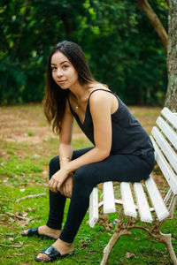 Full length portrait of young woman sitting on bench at field