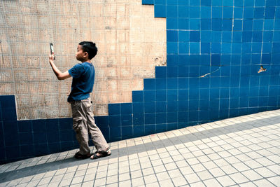 Full length of boy taking selfie through mobile phone while standing on footpath against tile wall