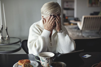 Worried senior woman with head in hands sitting by dining table at home