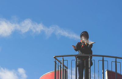 Low angle view of mature woman holding snow while standing at observation point against blue sky