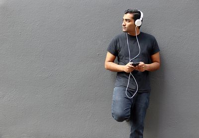 Young man looking away while listening music against wall