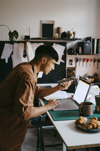 Side view of male freelancer doing multi-tasking with smart phone and laptop while working from home
