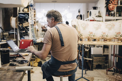 Rear view of male upholstery worker using laptop at workbench in workshop