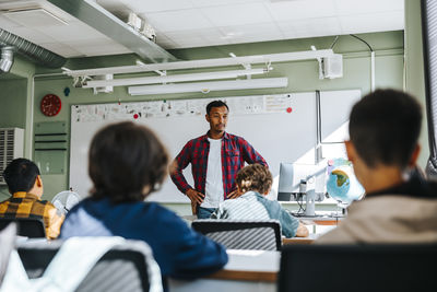 Male teacher with arms akimbo teaching group of students sitting in classroom at school