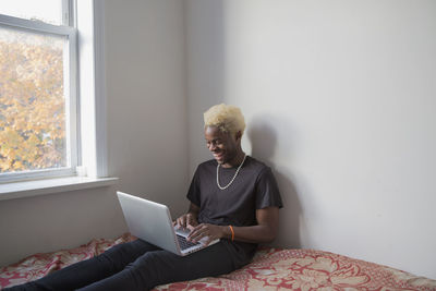Smiling young man using laptop while sitting on his bed