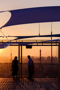 Rear view of silhouette man standing by railing against sky during sunset