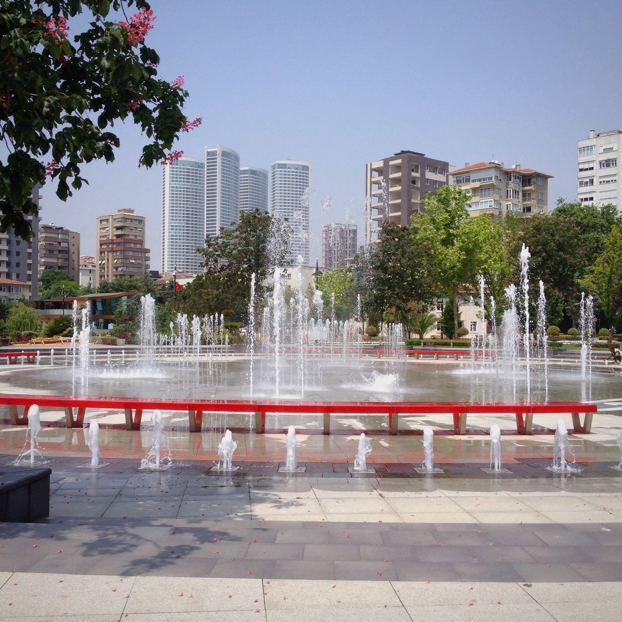 building exterior, architecture, built structure, city, tree, clear sky, modern, skyscraper, railing, office building, city life, sunlight, water, sky, building, cityscape, outdoors, day, sidewalk, incidental people