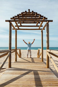 Back view of happy tourist standing with raised arms and barefoot on wooden boardwalk on sandy shore near sea on sunny day in summer