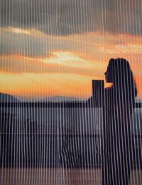 Woman standing by railing against sky at sunset reflecting on window