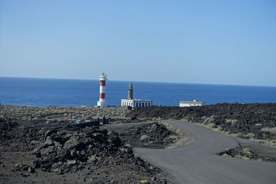 Lighthouse by sea against clear blue sky