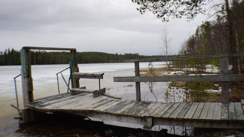 Empty bench on pier by lake against sky