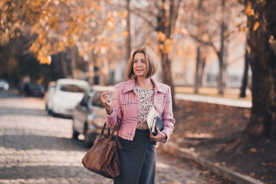 Happy business woman holding notebook and bag wearing stylish suit walking outdoors over leaves.
