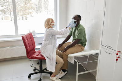 Doctor examining patient wearing protective face mask sitting on bed in clinic