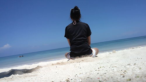 Rear view of woman sitting at beach against blue sky during sunny day