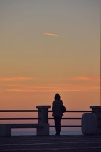 Rear view of silhouette woman standing on railing against sky during sunset