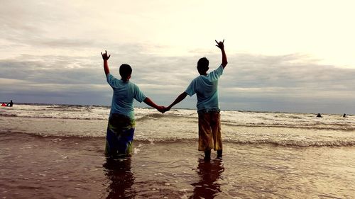 Rear view of boys holding hands while gesturing horn sign at beach