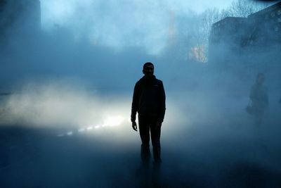 Silhouette man standing on street amidst smog at dusk