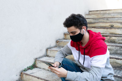 Young caucasian man wearing a face mask, sitting on some stairs, with earbuds, checking his phone