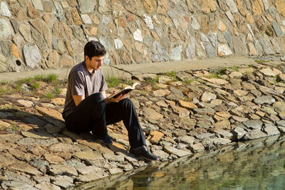 Full length of young man sitting on rock against wall