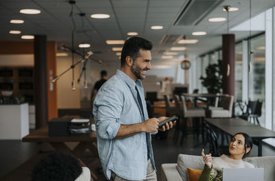 Side view of happy mature businessman standing with smart phone by female colleague during meeting