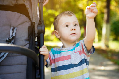 Pensive toddler boy stands near the stroller in the park with his hand raised.