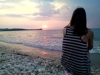 Rear view of woman sitting at beach against sky during sunset