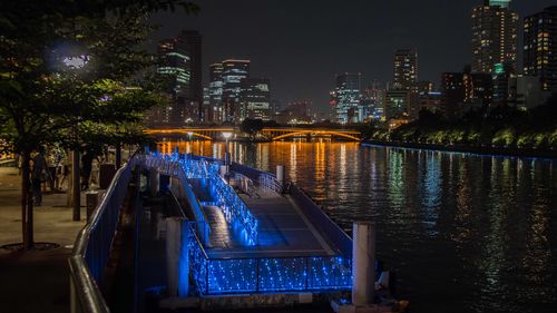 Illuminated modern buildings by river against sky at night