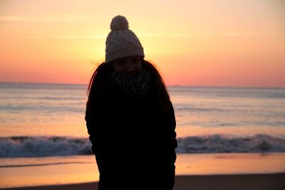 Portrait of young woman in warm clothing standing at beach against sky during sunset