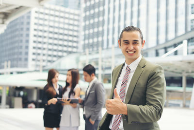 Portrait of smiling businessman gesturing against colleagues discussing on footpath 