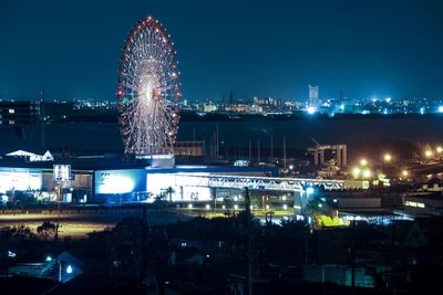 Illuminated ferris wheel by buildings against sky at night