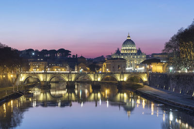 Illuminated buildings by river against sky at dusk