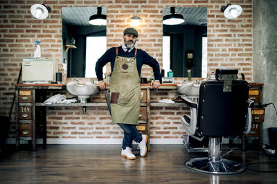 Full body of middle aged male barber in cap and apron standing near chair in grooming salon and looking at camera
