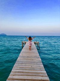 Rear view of woman with arms raised standing on pier over sea against sky