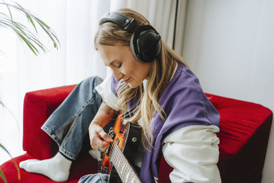 Guitarist wearing wireless headphones playing guitar on armchair