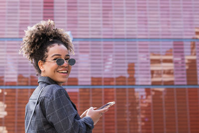 Happy african american female with curly hair and in casual outfit writing on smartphone in city and looking at camera