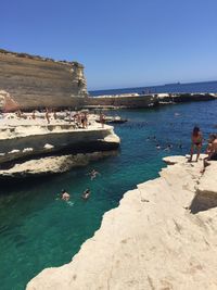 People on rocks by sea against clear blue sky