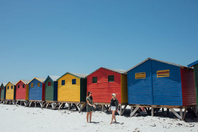 Friends at beach against colorful huts and clear blue sky during sunny day