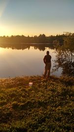 Rear view of man standing by lake against sky during sunset