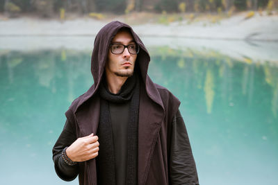 Bearded man in casual clothes with hood and glasses stands on shore