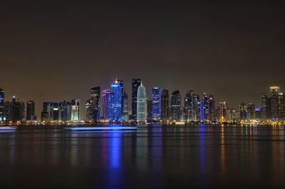 Illuminated modern buildings by bay against sky at night
