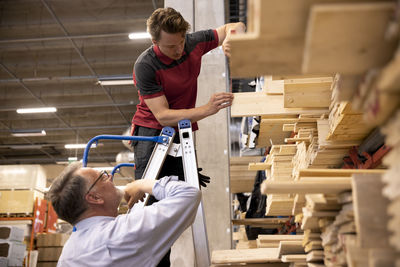 Salesman standing on ladder removing plank for male customer at hardware store